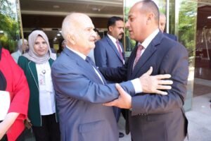 Two men in business suits warmly greet each other with a handshake outside a glass-building entrance, surrounded by colleagues