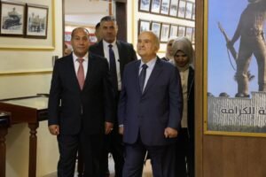Three men in suits walk through a photo-gallery hallway, with a woman in a hijab behind them and a large poster on the right.
