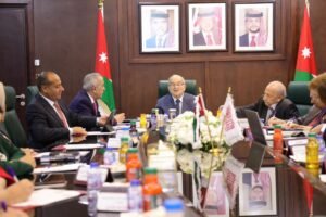 Group of officials in a formal boardroom meeting around a long table with Jordanian flags and framed portraits behind them as they discuss documents and laptops.