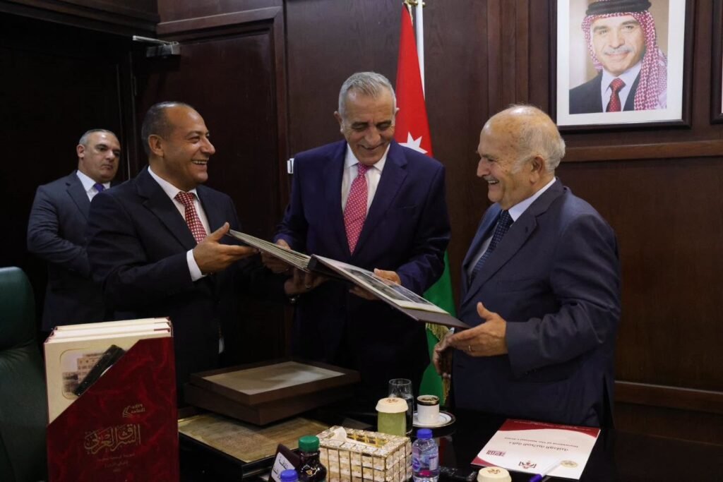 Three men in suits smile and exchange a large photo album in a wood-paneled office, with a flag and framed portrait in the background.