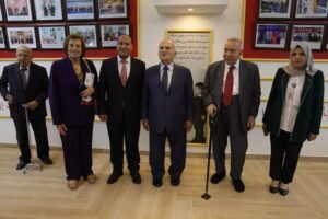 Six adults in formal attire stand together in a hallway with framed photos on the wall, posing for a group portrait in a gallery/event space.