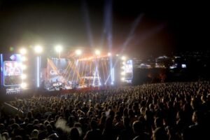 Outdoor nighttime concert with a large audience facing a brightly lit stage and tall screens on each side.