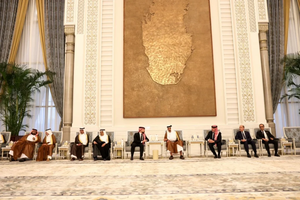 Formal meeting in an ornate hall with men in traditional Arab attire seated along a long row of chairs beneath a large textured wall panel.
