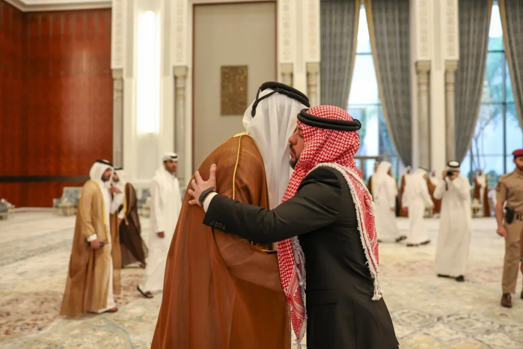 Two men in traditional Arab dress hug in a grand hall with tall curtains and ornate decor.",