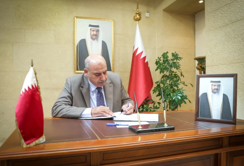 Bahrain official signing a document at a wooden desk, with the national flag and a framed portrait nearby.