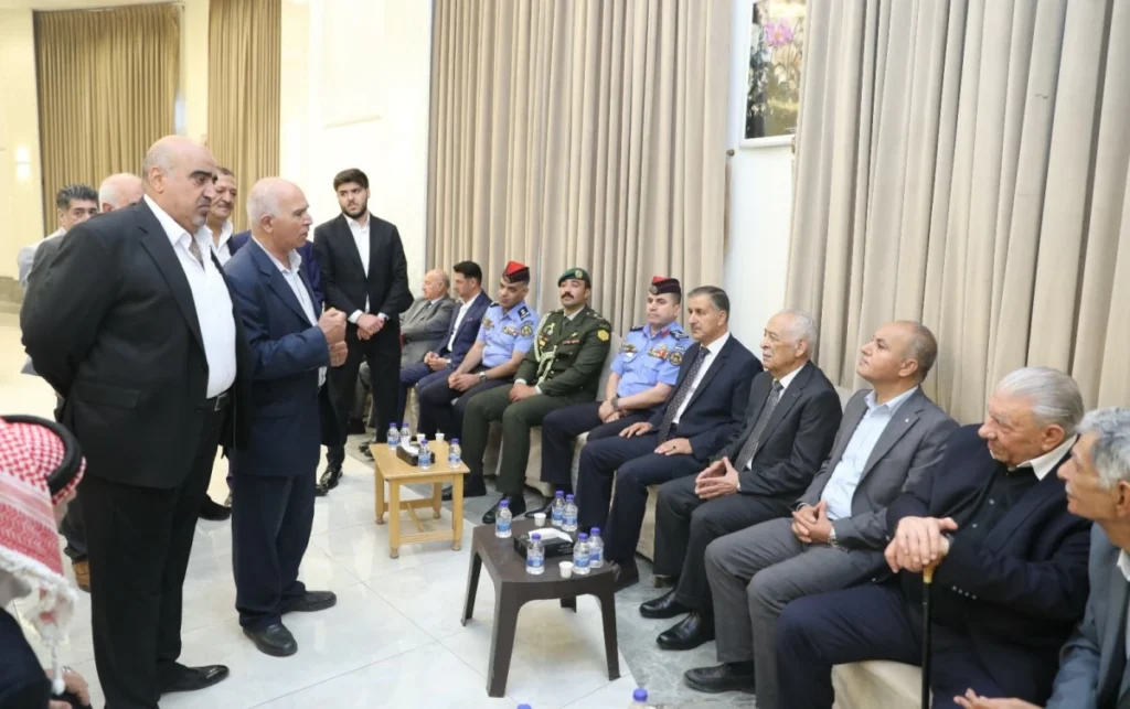 A group of men in suits and military uniforms seated along a wall while a man speaks to them in a formal indoor setting.