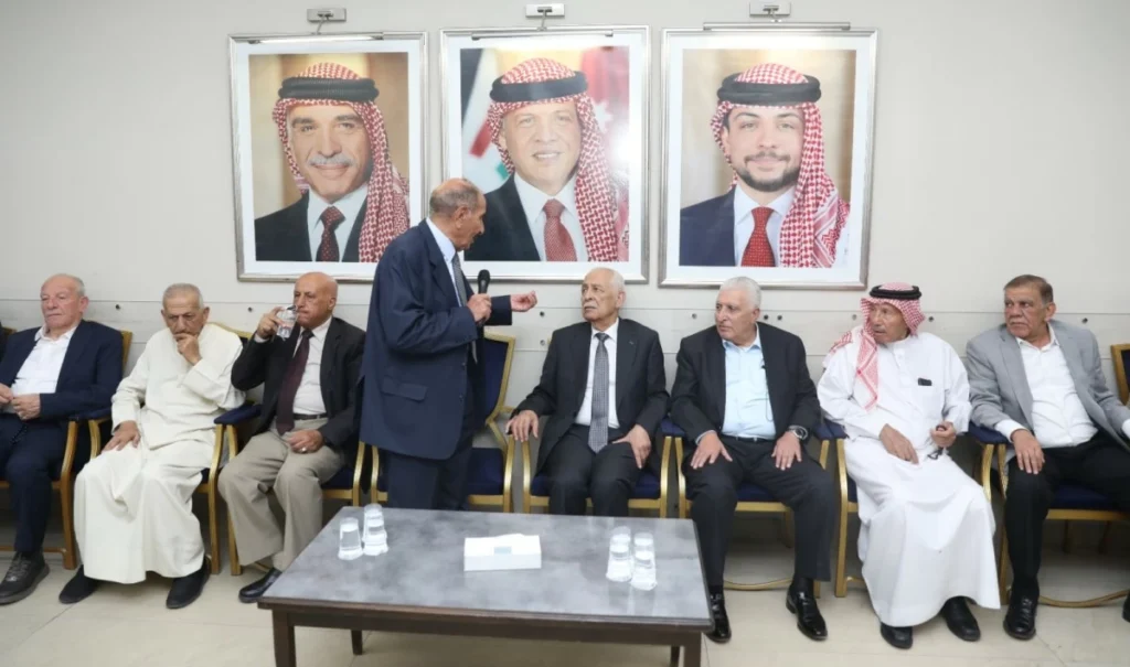 Nine men in suits or traditional thobes sit in a row in a conference room, one standing and speaking into a microphone.