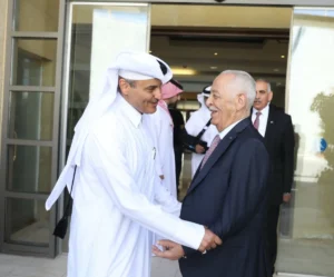 Two men greet each other with a handshake outside a building; one wears a white thobe and headscarf, the other a dark suit.”] ,