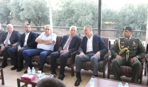 Group of men in suits sit in a row on a covered patio, with a uniformed soldier on the right.