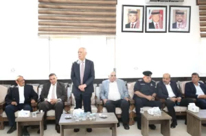 Group of men in suits seated on a sofa, one man standing and addressing the room, with three framed portraits on the wall behind.