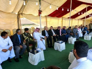 Group of men in traditional Arab dress and suits seated in a row under a striped tent at a formal meeting, with small tables of drinks in front.