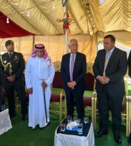 Group of men in a tent during a formal ceremony; an elderly man in white thobe holds a cane while others in suits stand nearby.