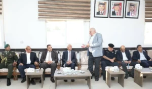 A group of men in suits and military uniforms sit on a couch as a man in a light gray blazer speaks standing beside them in a meeting room.