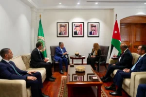Formal diplomatic meeting in a bright office: six suited officials seated on sofas around a central wooden coffee table, Jordan flag and another flag in the room, framed portraits on the wall behind them, a woman and five men engaged in discussion.