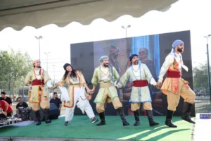 755890 - عروبة الاخباري | Oroba News Group of performers in traditional Middle Eastern costumes dance in a line on an outdoor stage, holding hands with a large screen backdrop behind them.