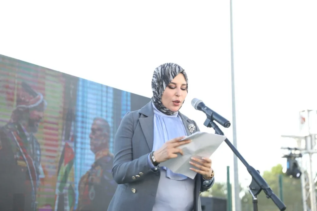 755893 - عروبة الاخباري | Oroba News Woman in hijab delivering a speech at a microphone on stage, reading from papers.