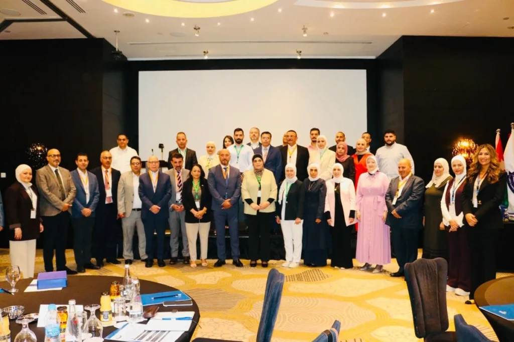 756086 - عروبة الاخباري | Oroba News Group of professionals posing for a conference photo in a ballroom, round tables with bottles in foreground.
