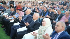 756093 - عروبة الاخباري | Oroba News Large audience seated outdoors, many clapping at a formal event; front row includes suited men and a woman in a hijab.