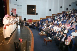 756330 - عروبة الاخباري | Oroba News Group of men in traditional white thobes and red-and-white keffiyehs holding swords on a stage, facing a seated audience in a theater venue.