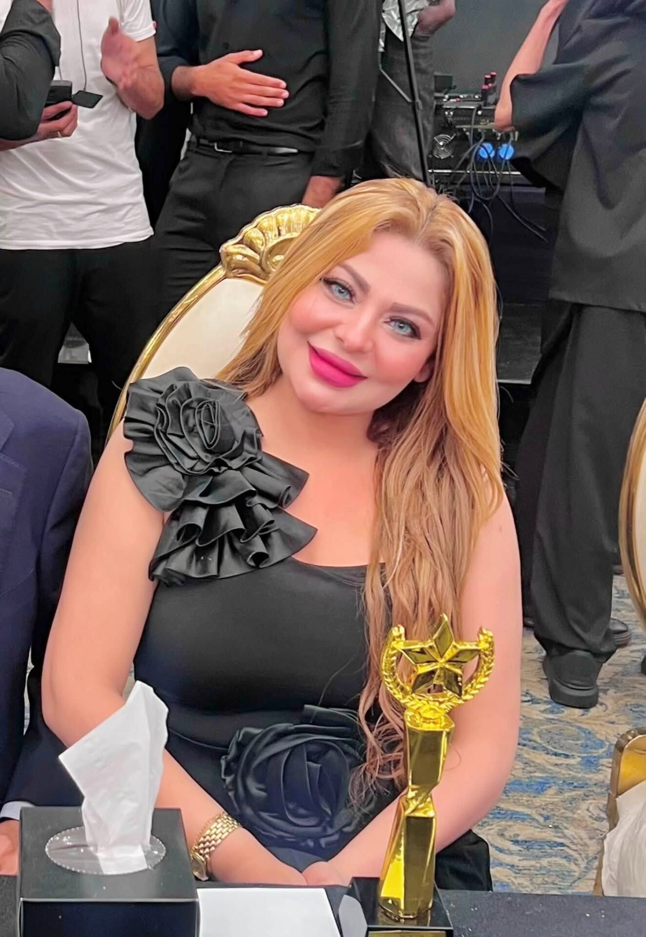 756436682_10243304403407765_8405866757271375939_n Smiling woman in a black dress with ruffled shoulder holding a gold trophy, seated at a table with tissues and awards nearby.