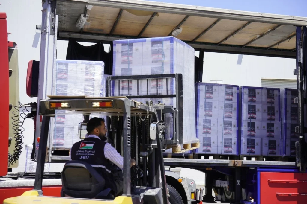 756470 - عروبة الاخباري | Oroba News Forklift operator in a reflective vest drives a forklift toward stacked, wrapped pallets on a truck at a loading area.