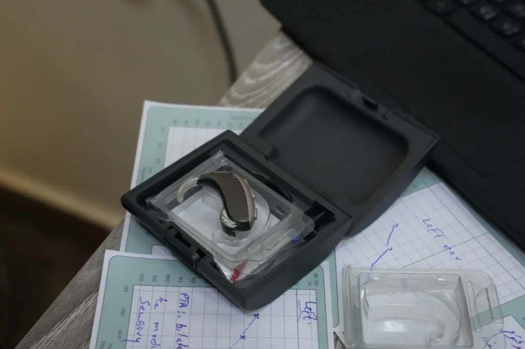 756471 - عروبة الاخباري | Oroba News Hearing aid in a clear case on a desk next to a laptop and handwritten notes on graph paper.