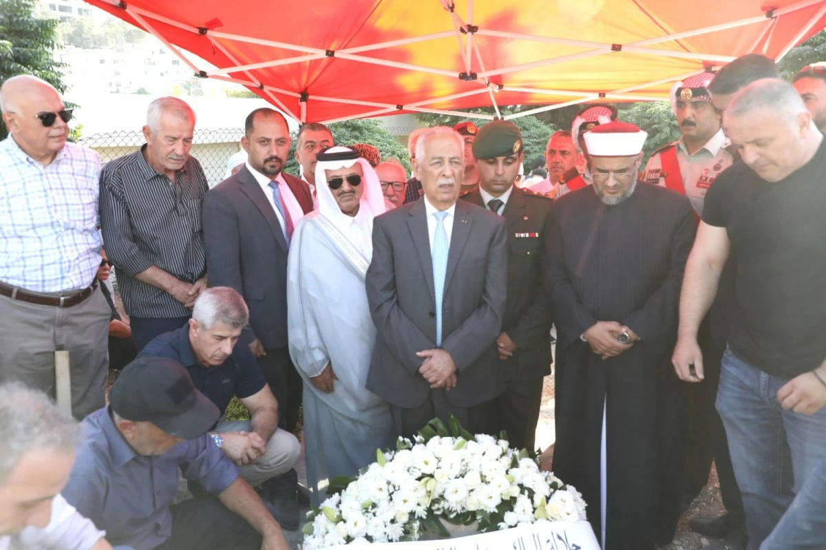 756887 A group of men in suits, traditional robes, and a uniformed officer stand under a red canopy at a graveside funeral, near a white flower wreath.