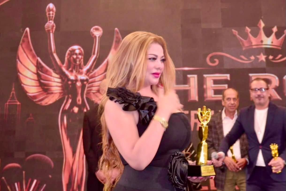 757937320_10243304400927703_1277102467234886323_n Woman in a black dress holding a gold trophy on stage at an award ceremony, with a red backdrop and other people in the background.