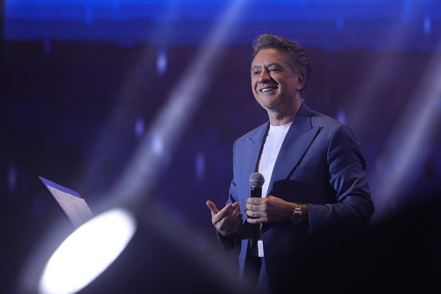 758193311_1010858648226902_5997663496886078745_n Male presenter in a blue suit on stage, smiling, holding a microphone under blue spotlight.