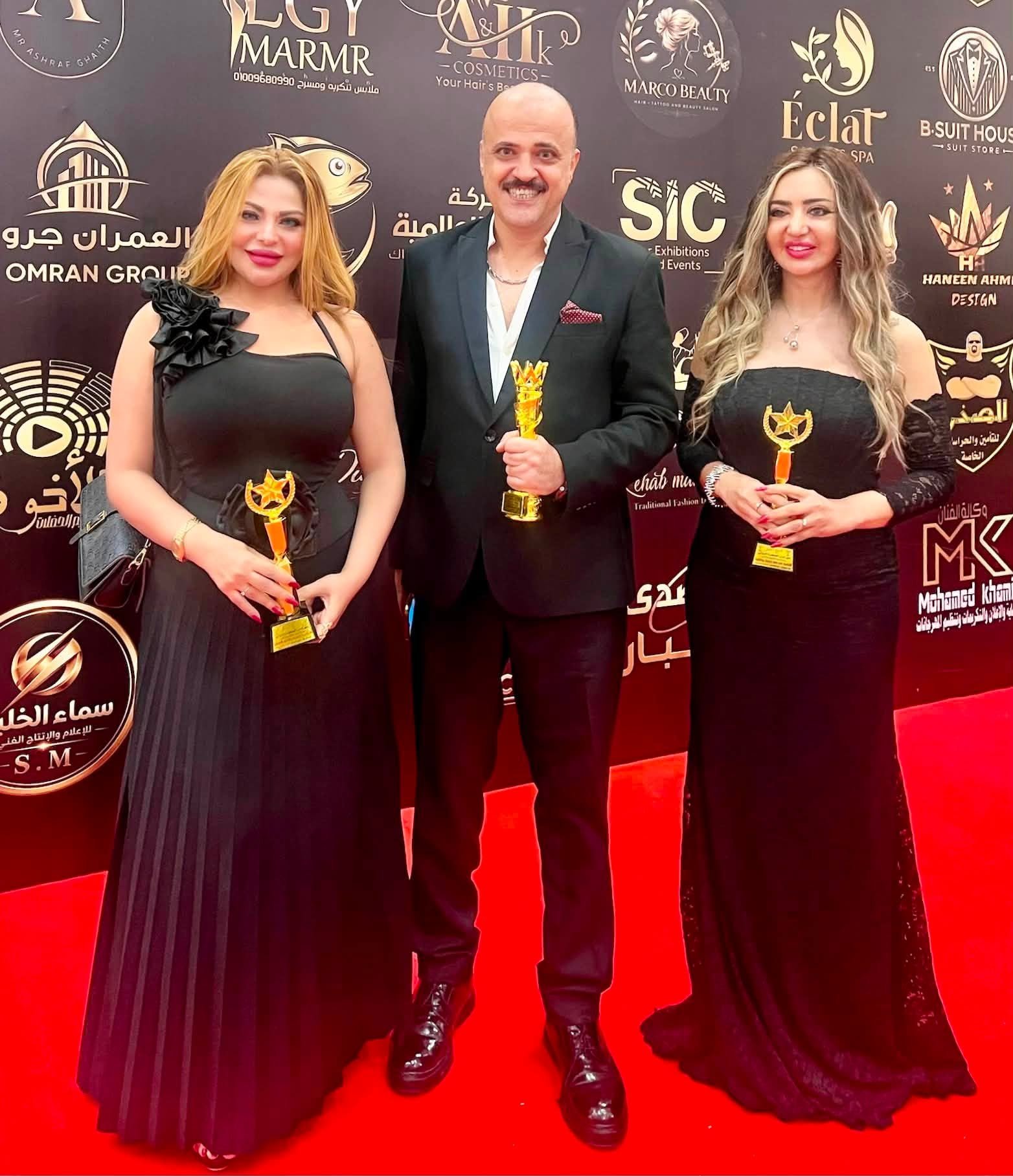 758542834_10243304403327763_89607770437605201_n Three people in formal attire on a red carpet, each holding a gold trophy at an awards backdrop.