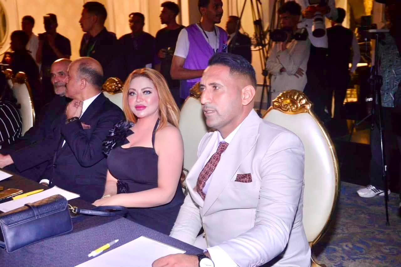 758617370_10243304398047631_2512642080702554600_n Man in a light gray suit and patterned tie sits beside a woman in a black dress at a formal event, with photographers in the background.