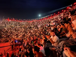 Large night-time amphitheater filled with cheering spectators under red lights.