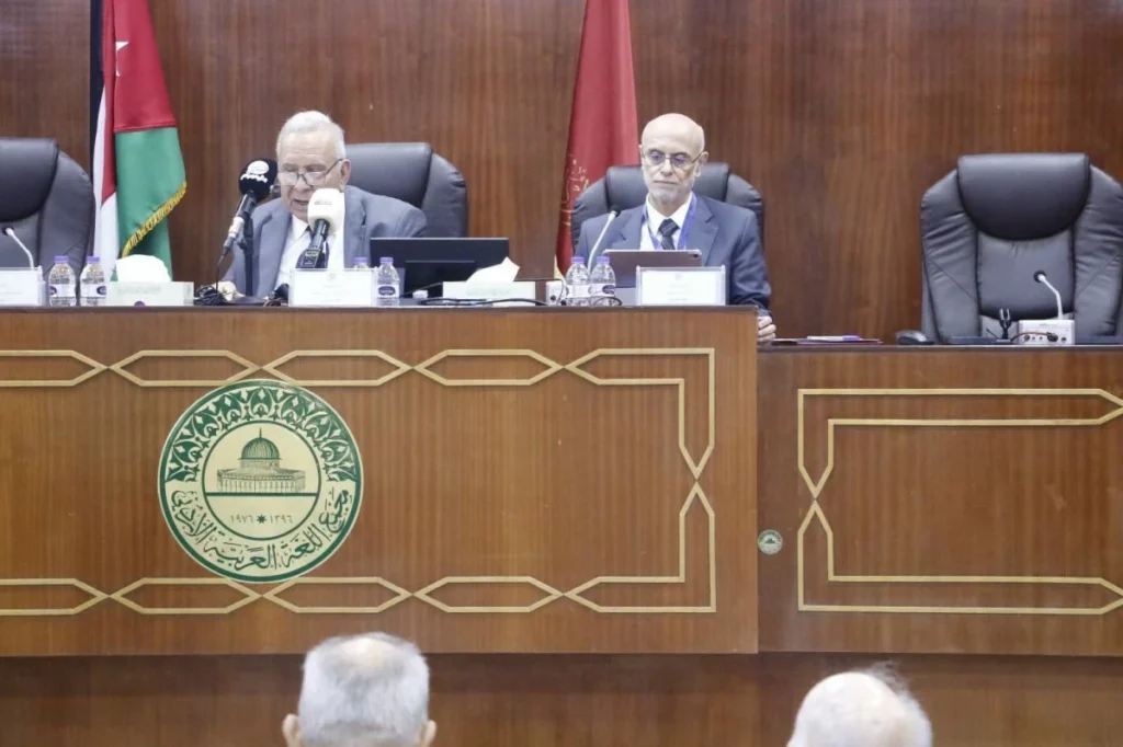 Two men in suits sit at a wooden conference desk with microphones and water bottles, an emblem on the front, and flags behind them.