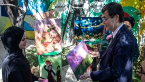 Man in a suit and a woman in a headscarf exchange a painting in a sunny courtyard filled with colorful murals and art pieces nearby.