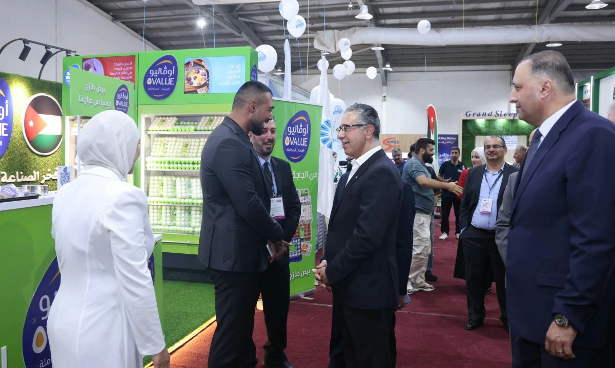 759178 Businesspeople greet each other with a handshake at an expo booth in a green-themed display area, while a woman in white looks on.