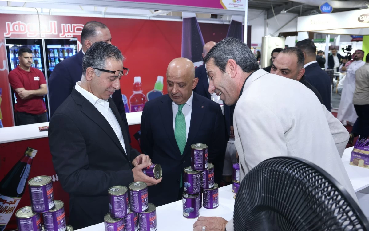 759180 Three men in suits inspect purple-labeled cans at a trade show booth, with a red backdrop and display stacks on the counter.