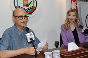 Man speaking at a press conference desk with a microphone, wearing glasses and a blue shirt; woman in a purple blazer sits beside him, logo on wall behind them.