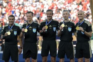 FBL-WC-2026-MATCH104-ESP-ARG-FINAL-TROPHY - عروبة الاخباري | Oroba News Five FIFA referees in black uniforms posing with gold medals after a ceremony, stadium crowd in the background.