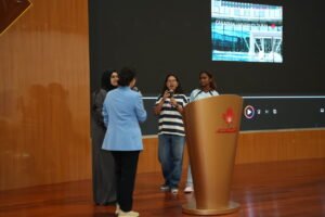 DSC07057 - عروبة الاخباري | Oroba News Student speaks into a microphone at a large golden podium on stage, flanked by two peers, with a university screen in the background