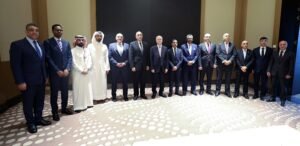 IMG_1684 - عروبة الاخباري | Oroba News Group of men in suits (and a couple in traditional thobes) posing for a formal line photo in a conference setting with a patterned carpet behind them.