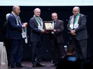 Four men in suits on a stage; a plaque is being handed to the smiling recipient between two presenters wearing scarves.