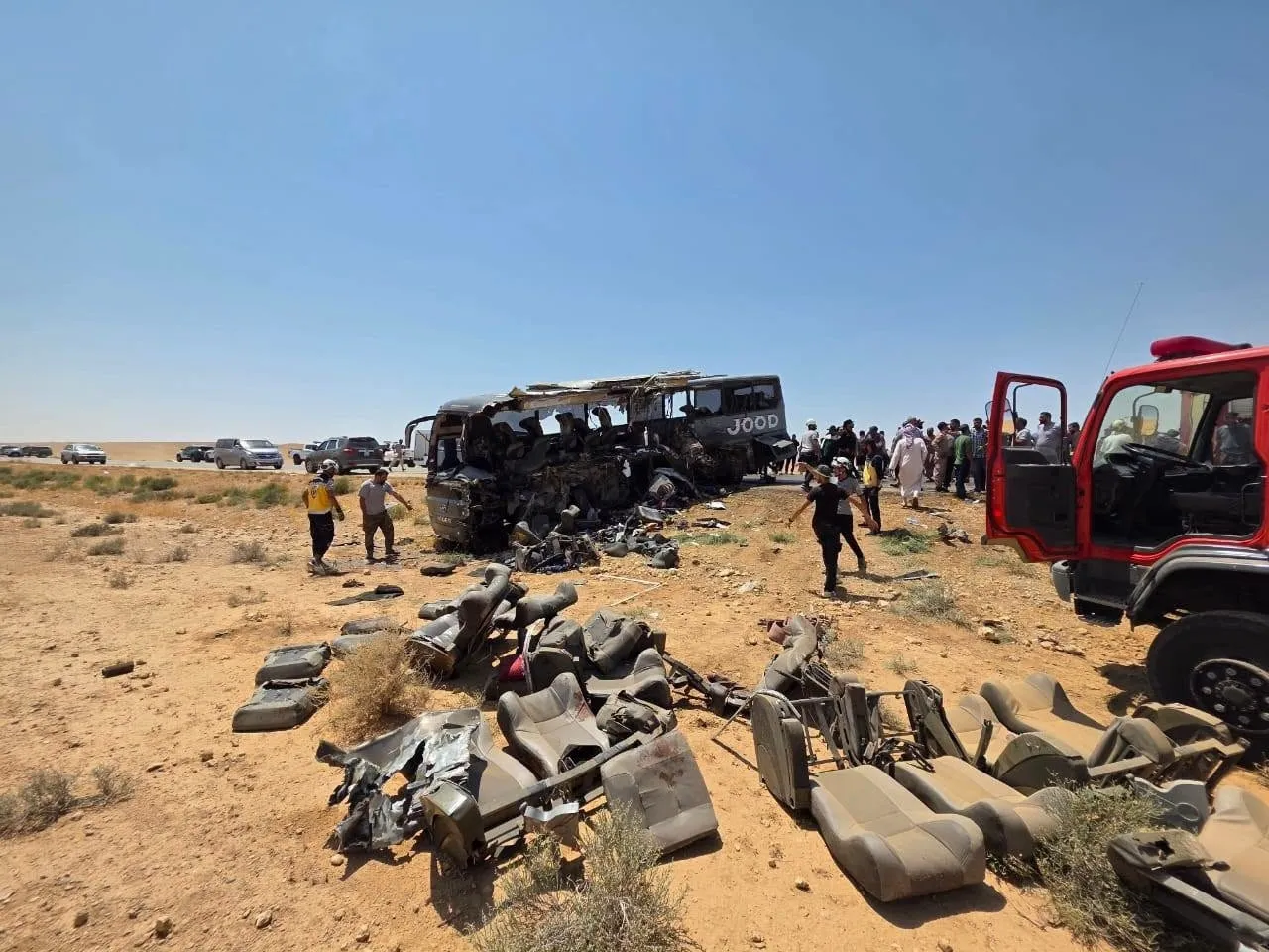 Image3_7202625141458570913050 Desert highway crash scene with a heavily damaged bus, scattered seats and debris, onlookers and emergency vehicles nearby.