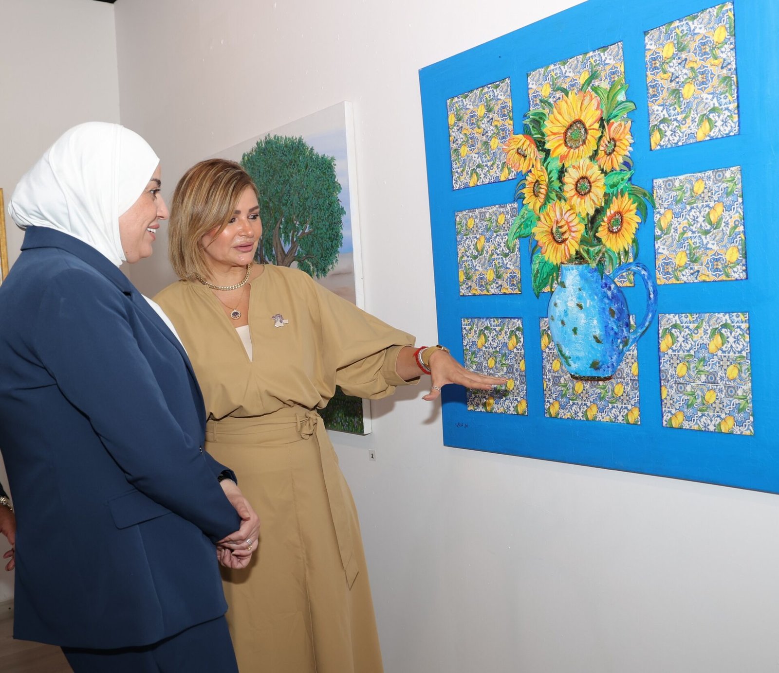 M3Afigz5CE2HFDwYIxLAtVDWgUjEg0x31f4EDL8Z Two women in professional attire view a large sunflower painting mounted on a bright blue wall; one woman in beige points at the artwork while the other smiles in a navy outfit.
