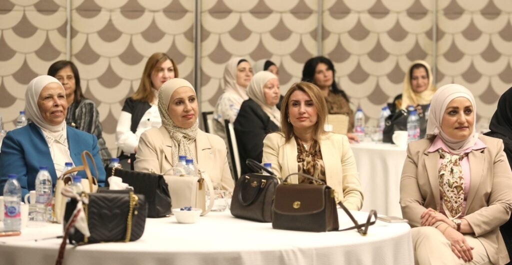 MgxrQeaPRjxXymaSR3G4TNrrwvoe2cWlJXzxwCkI - عروبة الاخباري | Oroba News Group of professional women seated around a table at a conference, with handbags and water bottles visible on the table.