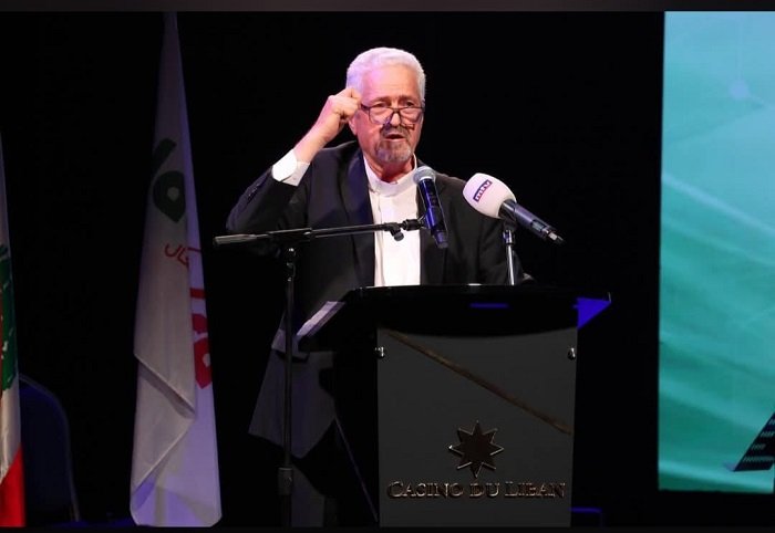 WhatsApp Image 2026-07-05 at 104000 AM - عروبة الاخباري | Oroba News Older man in a dark suit speaks at a podium with two microphones on a stage on a conference backdrop.