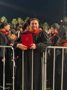 Graduate in cap and gown holding a red diploma, smiling at a nighttime graduation ceremony.