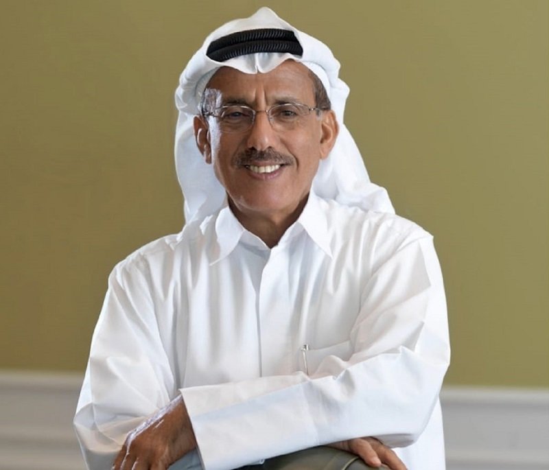 Smiling man in a white thobe and headscarf (ghutra) posing with arms crossed against a neutral wall background, indoors.