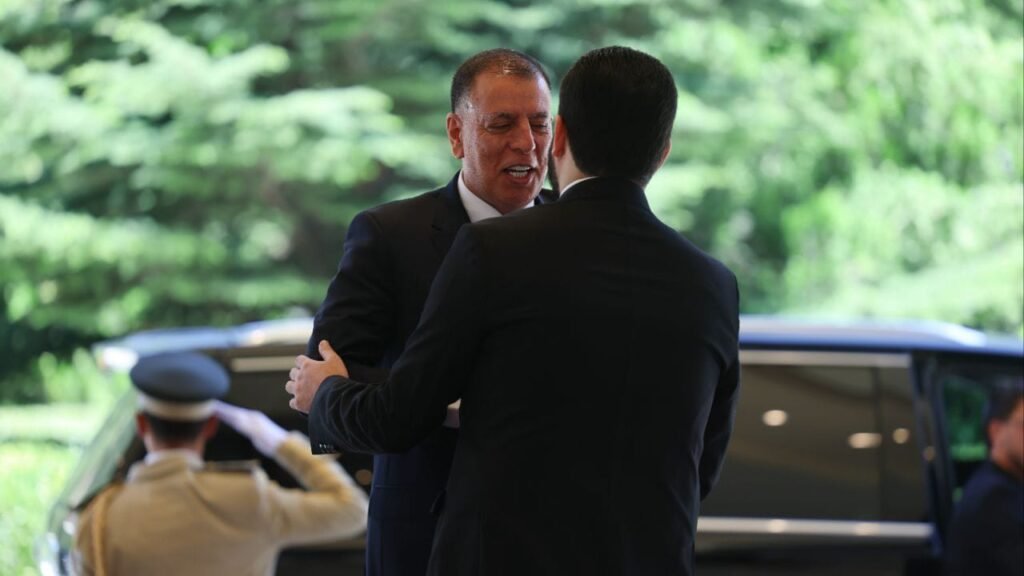 Two men in business suits shake hands outside beside a dark SUV, with a uniformed officer saluting in the background and greenery behind them.