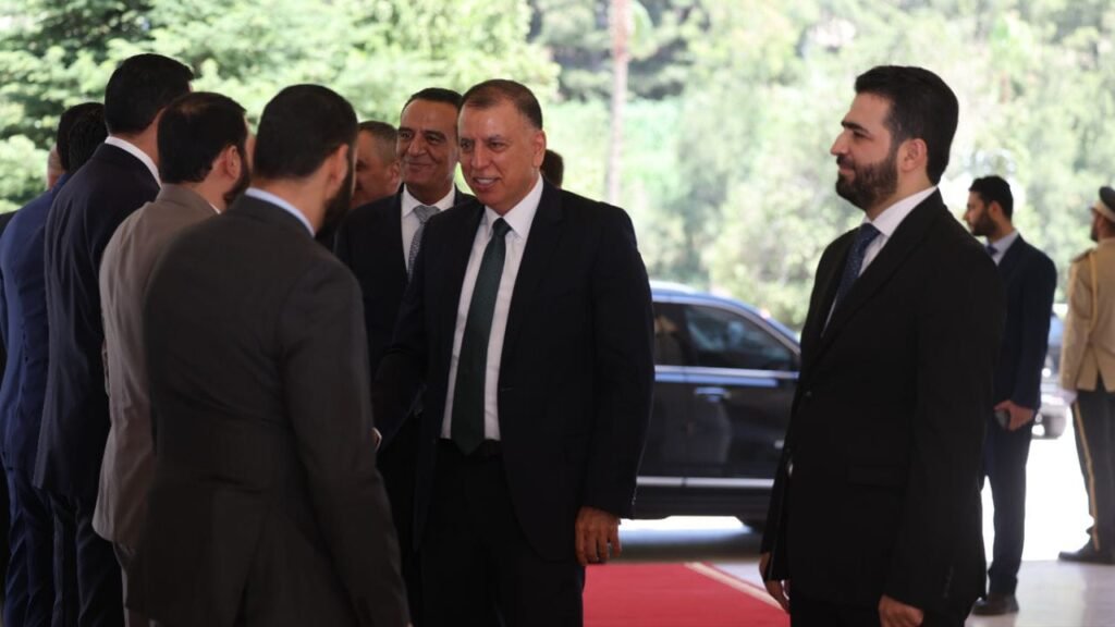 Group of formally dressed men greeting each other and shaking hands on a red carpet outdoors, with a car and greenery in the background.