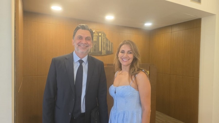 Smiling man in a dark suit and a woman in a light blue dress pose together in a warmly lit hallway or lobby area.